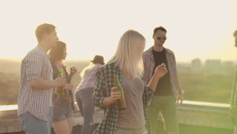 The-American-blonde-woman-is-dancing-on-the-roof-with-her-friends-who-drinks-beer-on-the-party.-She-smiles-and-enjoys-the-time-she-is-dancing-with-a-beer-in-a-gray-tank-top-and-a-dark-green-plaid-shirt.